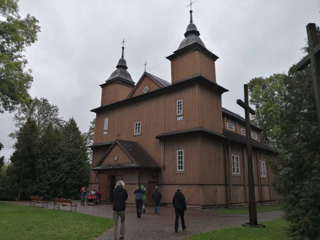 Drewniany kościół, przed grupa osób; pogoda deszczowa