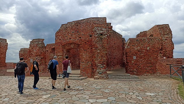 ceglane ruiny, przed nimi 4 idących mężczyzn, odwróconych przodem do pozostałości budowli